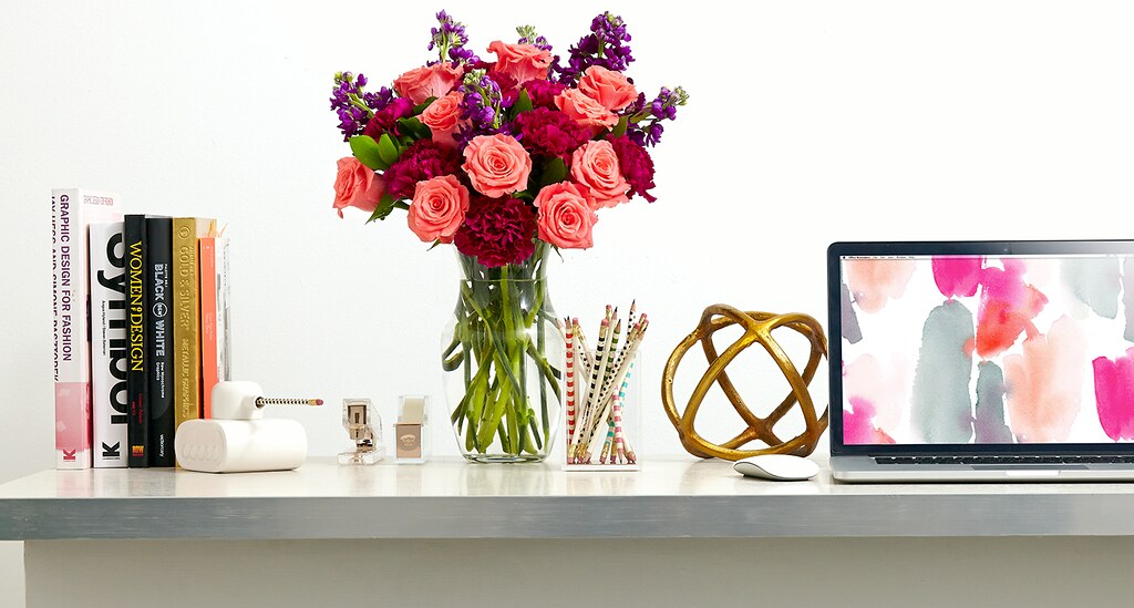 pink and red carnations and roses in a glass vase on a desk with laptop computer pencils books stapler tape roll tank