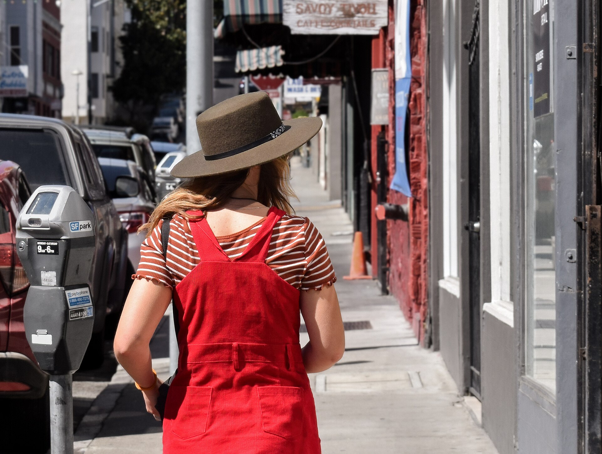 A woman wearing a wide-brimmed hat and a red dress walks down a sidewalk in the North Beach neighborhood of San Francisco, California