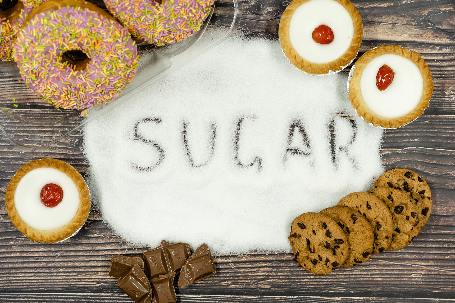 It might look tempting to some. But the sugars in common junk foods like doughtnuts, cakes, chocolate and cookies is immense. Sugar is incredibly damaging when consumed in high quantities. IMage depicts the word "sugar" written out in sugar with a series of sugary snacks surrounding. Image created by Formulate Health and distributed under Creative Commons. We welcome you to use commercially or non commercially with attribution in the form of a link to the creator at formulatehealth.com/